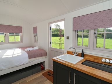 A small room with a bed with folded towels a sink and cooktop under windows with patterned blinds at Dragonfly Shepherd's Hut in Oakford near Aberaeron