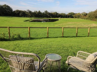 Two outdoor chairs and a small round table on grass facing a wooden fence and a grassy field with a pond beyond at Dragonfly Shepherd's Hut in Oakford near Aberaeron