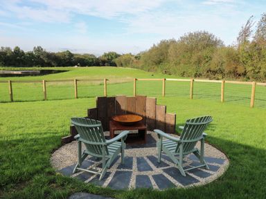 Outdoor fire pit area with two green wooden chairs and wooden windbreak in a grassy field at Dragonfly Shepherd's Hut in Oakford near Aberaeron