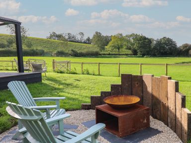 An outdoor seating area with wooden chairs around a metal fire pit and a wooden fence in a grassy field at Dragonfly Shepherd's Hut in Oakford near Aberaeron