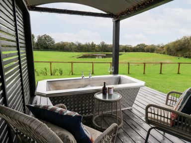 An outdoor covered patio with a metal bathtub filling with water a round glass table with bottles and two wicker chairs overlooking a green field