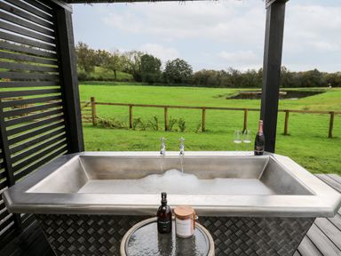 An outdoor metal bathtub with running water soap and bath salts on a small table and a bottle of champagne with glasses overlooking a green field at Dragonfly Shepherd's Hut Oakford near Aberaeron