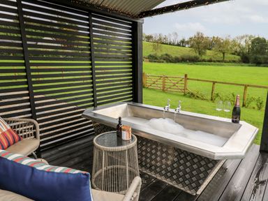An outdoor covered patio with a metal bathtub filled with bubbles next to a small round table and seating overlooking a green field at Dragonfly Shepherd's Hut in Oakford near Aberaeron