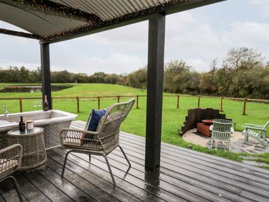 A covered porch with chairs and a small table by a metal tub overlooking a fenced grassy yard with a fire pit and chairs at Dragonfly Shepherd's Hut in Oakford near Aberaeron