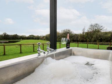 A filled outdoor metal bathtub with running water two glasses and a bottle of champagne on the edge overlooking a fenced grassy field at Dragonfly Shepherd's Hut in Oakford near Aberaeron