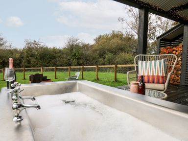 An outdoor area with a bubble bath tub champagne glasses and bottle a wicker chair with patterned cushion stacked firewood and green grass beyond a wooden fence at Dragonfly Shepherd's Hut in Oakford near Aberaeron