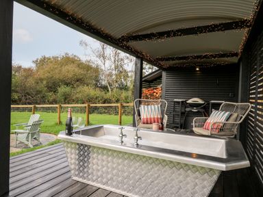 An outdoor covered patio with a metal bathtub two chairs and a grill with grass and trees in the background at Dragonfly Shepherd's Hut Oakford near Aberaeron