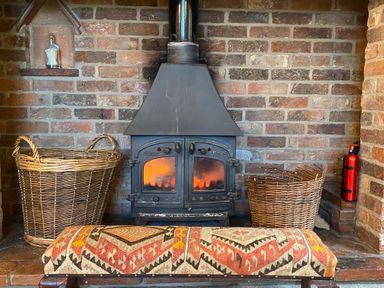 A wood-burning stove with fires inside flanked by wicker baskets and a patterned footstool in front at Rivendell in Weybread near Harleston