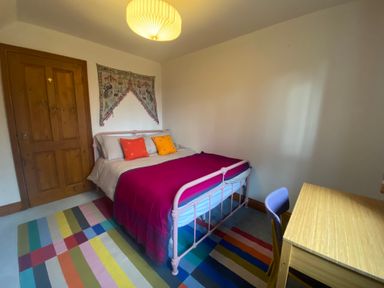 A bedroom with a bed colorful cushions a striped rug a wooden desk and chair at Rivendell in Weybread near Harleston