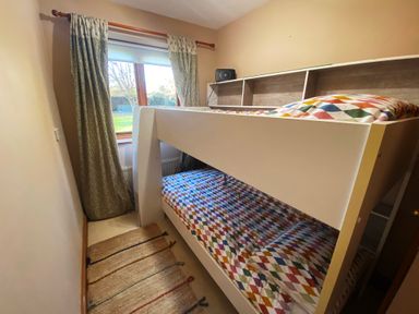 A small bedroom with a white bunk bed and patterned bedding next to a window with curtains and a rug on the floor at Rivendell in Weybread near Harleston