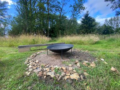 An outdoor fire pit surrounded by rocks with a wooden bench nearby in a grassy area with trees in the background at Rivendell in Weybread near Harleston