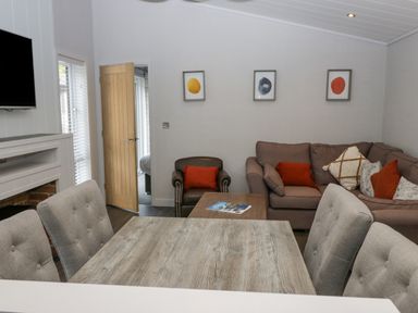 A living room with a wooden dining table gray chairs a brown sofa orange cushions a leather armchair three framed prints on the wall and a TV above a fireplace at The Bothy in Keswick