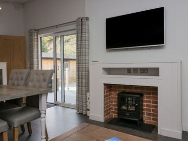 A dining area with upholstered chairs and a wooden table next to a sliding glass door and a fireplace with a mounted TV at The Bothy in Keswick