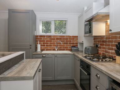 A kitchen with grey cabinets brick backsplash gas stove toaster and a sink under a window at The Bothy in Keswick