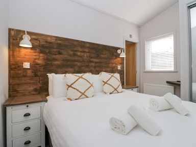 A bedroom with a bed, wooden headboard, bedside tables, wall lamps, pillows, rolled towels, window, and door at The Bothy in Keswick