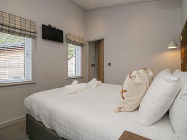 A bedroom with a large bed with pillows and rolled towels two windows with blinds and a mounted television at The Bothy in Keswick