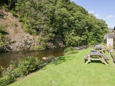A grassy riverbank with picnic tables and trees along the water at The Bothy in Keswick