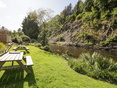 A grassy area with picnic tables next to a river and trees at The Bothy in Keswick
