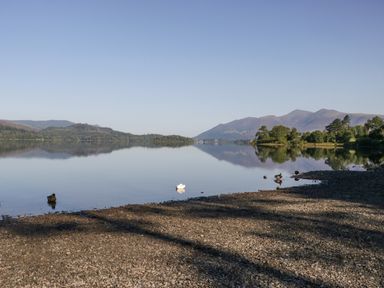 A calm lake with ducks on water and pebbled shore with mountains and trees in the background at The Bothy in Keswick