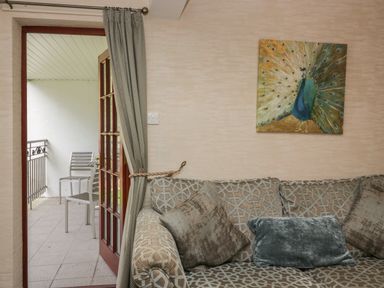 A sofa with cushions and a painting of a peacock above it next to a door opening to a balcony with two chairs at Apartment 1 in Keswick