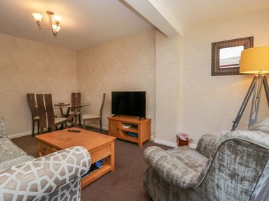 A living room with a patterned sofa and armchair a wooden coffee table a TV on a wooden stand a glass dining table with wooden chairs a floor lamp and a wall mirror at Apartment 1 in Keswick