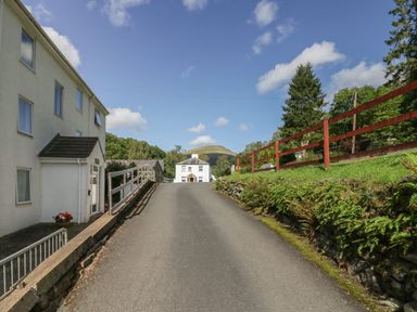 A paved driveway with a white house at the end surrounded by greenery and a red fence at Apartment 1 in Keswick