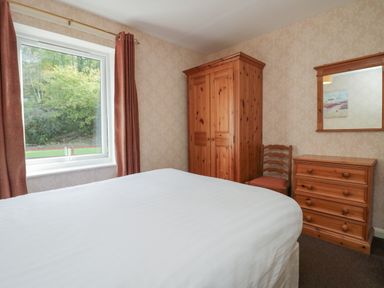 A bedroom with a bed a wooden wardrobe a wooden chest of drawers a wooden chair and a window with curtains at Apartment 2 in Keswick