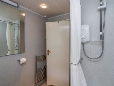 A bathroom with a shower, shower curtain, wall-mounted shower controls, a mirror, toilet paper holder, heated towel rail, and a door at Apartment 3 in Keswick