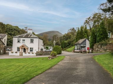A rural road with a white house and a small building surrounded by greenery and hills in the background at Apartment 3 in Keswick