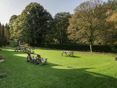 An outdoor grassy area with wooden picnic tables and chairs surrounded by trees at Apartment 4 in Keswick