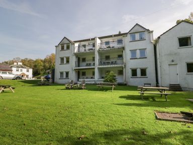 An outdoor lawn area with picnic tables and chairs next to a three-story white building with balconies at Apartment 4 in Keswick