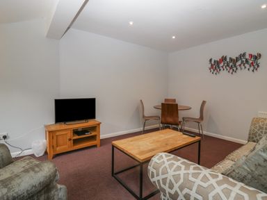 A living room with a television on a wooden stand a wooden coffee table a round dining table with four chairs and wall art at Apartment 6 in Keswick