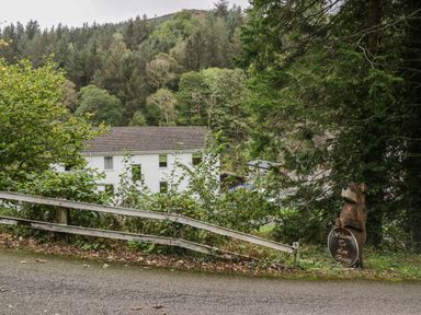 A wooded hillside with a white building and a wooden bear statue holding a welcome sign near a road at Apartment 6 in Keswick