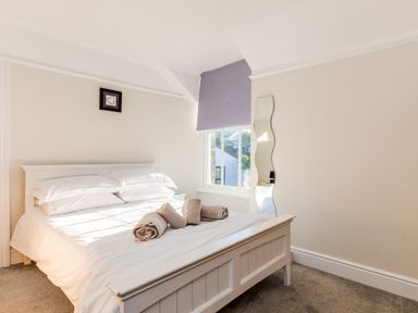 A bedroom with a white bed, rolled towels on the bed, a wavy mirror on the wall, and a window with a gray blind at Tre Lowen in Port Isaac