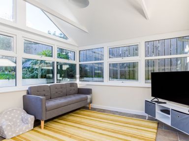 A room with large windows featuring a gray sofa pet chair a yellow striped rug and a TV on a white stand at Tre Lowen in Port Isaac