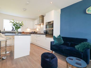 A kitchen with white cabinets and island next to a blue wall with a sofa and small table at Cobalt in St Eval near Mawgan Porth