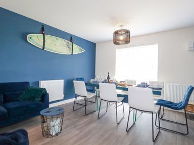 A dining room with a glass table set for a meal with white and blue chairs and a surfboard on a blue wall at Cobalt in St Eval near Mawgan Porth