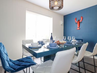 A dining room with a blue dining table set with plates glasses cutlery and a bottle with blue and white chairs and a red lobster wall decoration at Cobalt St Eval near Mawgan Porth