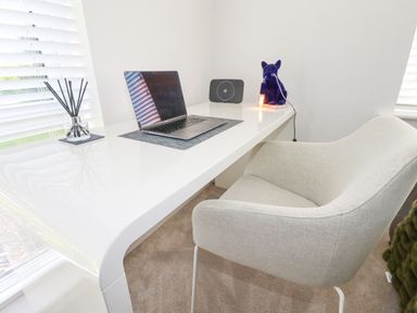 A white desk with a laptop diffuser speaker and a chair in a room with white walls and windows