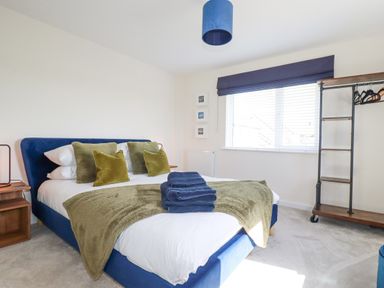 A bedroom with a blue bed, green pillows and blanket, wooden bedside table, open closet, and window with blinds and curtain at Cobalt in St Eval near Mawgan Porth