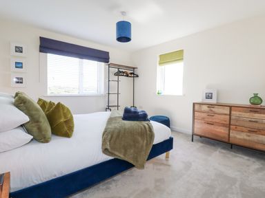 A bedroom with a bed with green pillows and blanket a wooden dresser a clothing rack and two windows with blinds at Cobalt in St Eval near Mawgan Porth
