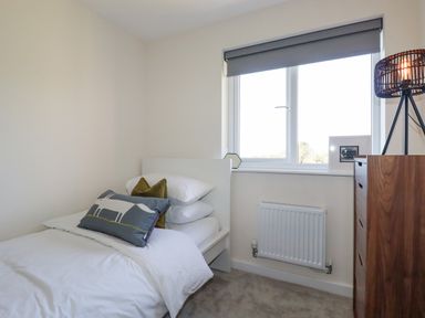 A small bedroom with a single bed white bedding pillows a window with a blind a radiator a wooden chest of drawers and a lamp at Cobalt in St Eval near Mawgan Porth
