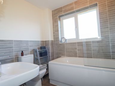 A bathroom with a bathtub under a window a toilet a washbasin and a small shelving unit with towels at Cobalt in St Eval near Mawgan Porth