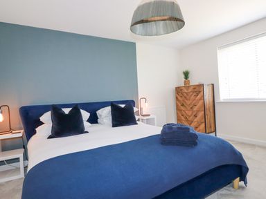 A bedroom with a bed with blue bedding two nightstands with lamps wooden chest of drawers and a window with blinds at Cobalt St Eval near Mawgan Porth