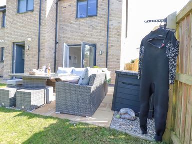 An outdoor patio area with grey wicker furniture and table next to a brick house with open doors and a black wetsuit hanging on a wooden fence at Cobalt in St Eval near Mawgan Porth