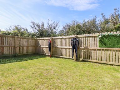A grassy backyard enclosed by a wooden fence with two wetsuits hanging on the fence at Cobalt in St Eval near Mawgan Porth
