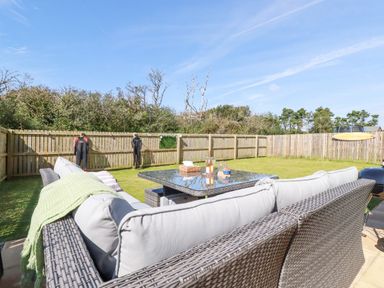 An outdoor seating area with a wicker sofa and glass table in a fenced grassy yard with wetsuits and a surfboard hanging on the fence at Cobalt in St Eval near Mawgan Porth
