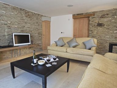 A living room with beige sofas a black coffee table with wine glasses and a bottle a television on a wall shelf and stone walls at The Milking Shed in Bantham