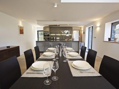 A dining room with a table set with plates and glasses in front of a kitchen at The Milking Shed in Bantham