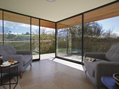 A corner seating area with two armchairs and small tables in a room with large glass walls at The Milking Shed in Bantham